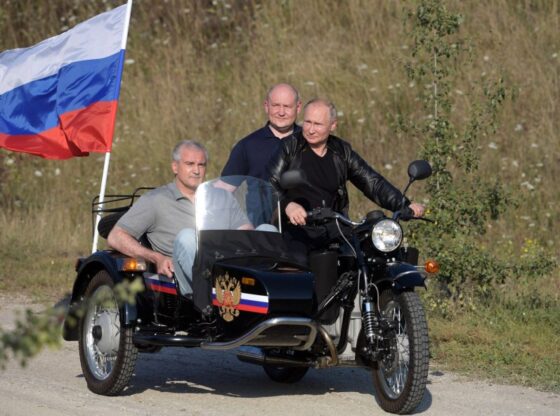 Vladimir Putin riding a motorcycle with a sidecar, accompanied by two men and a Russian flag.