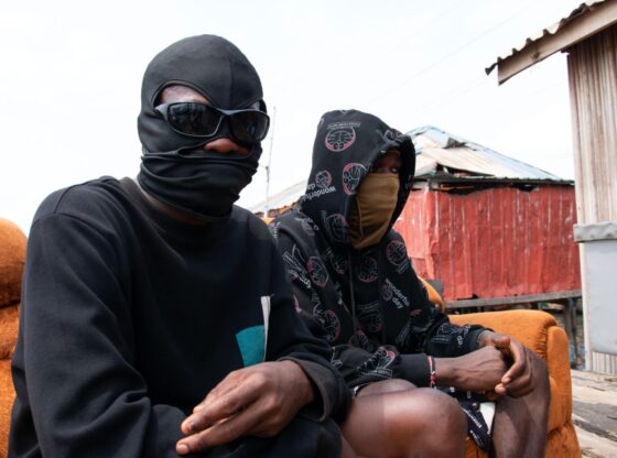 Two individuals in masks and hoodies sit on an orange couch outdoors.