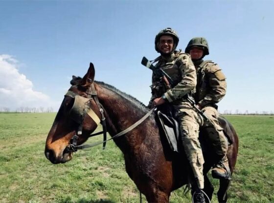 Two soldiers in camouflage and helmets ride a brown horse across a grassy field.