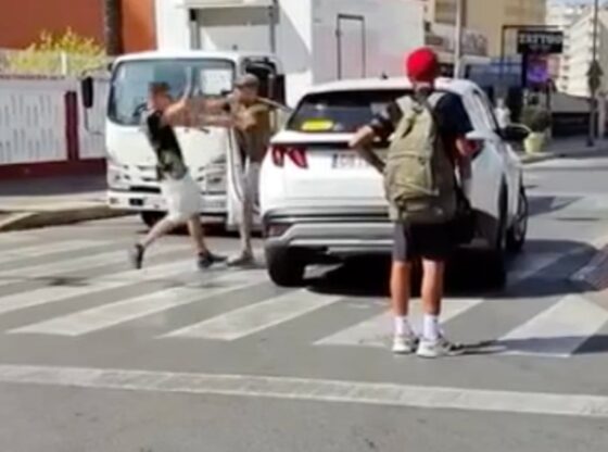 A man in a black shirt and white shorts argues with another man wearing a brown shirt and hat, who is standing next to a white car on a zebra crossing.